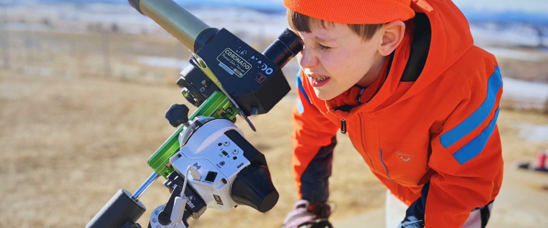 A student in an orange jacket looks through a solar telescope outdoors.