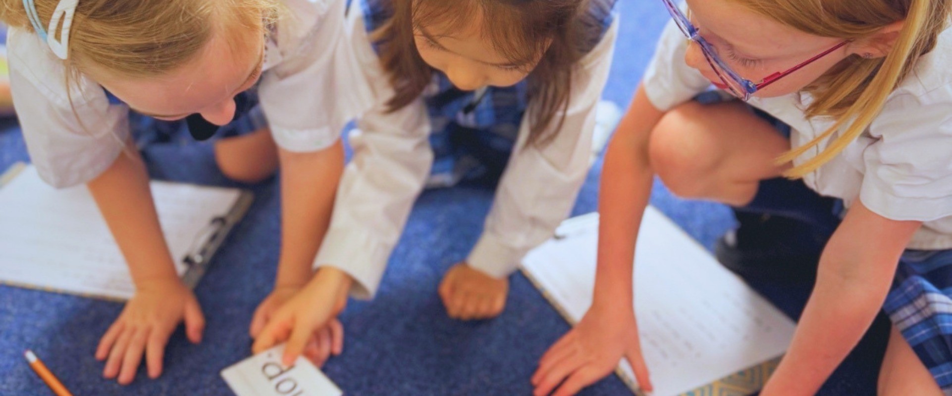 Three students kneel on the carpet, working with word cards and clipboards.