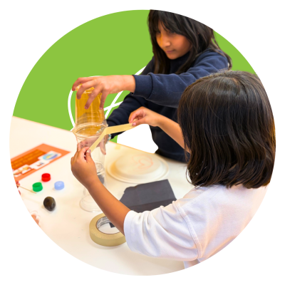 Two River Valley School students working together on a craft project with paper, sticks and tape, on a green circular background.