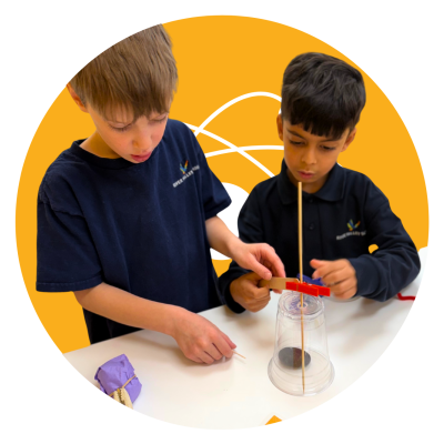 Two River Valley School students in uniform building a structure with a jar, sticks and elastic bands, on a yellow circular background.